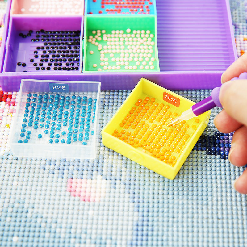 Yellow diamond painting tray filled with orange drills being picked up by a multi-tip pen, surrounded by color-coded trays – perfect for organized DIY creators.