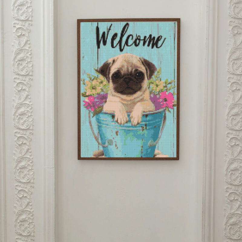 Framed diamond painting featuring a cute pug in a blue bucket with flowers, ideal for pet lovers seeking cheerful wall decor or a handmade welcome sign.