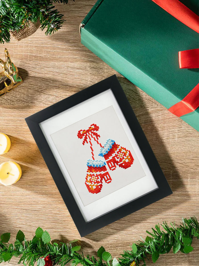 Framed mini diamond painting of red and blue Christmas mittens tied with a bow, placed beside holiday gift box and candles – beginner-friendly holiday diamond art for cozy seasonal crafting.