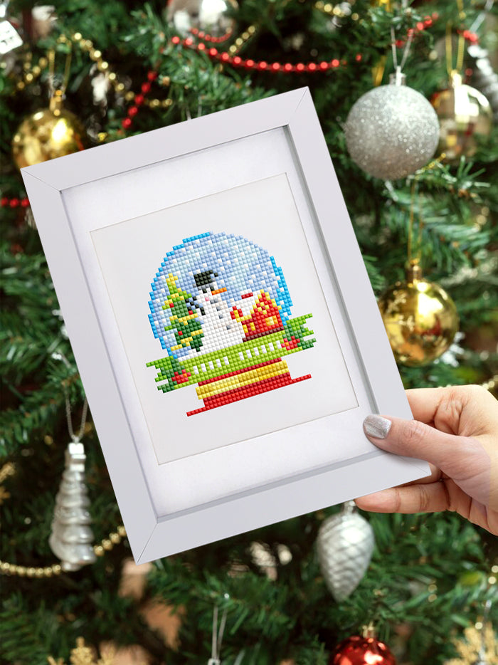 Diamond painting of a holiday snow globe framed on a tree with ornaments. Soft canvas and resin drills make it a beginner-friendly Christmas craft.